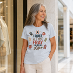 A smiling older woman stands near a boutique wearing a powder blue graphic T-shirt with the phrase 'I Quilt So I Don't Unravel in Real Life' and quilt-themed illustrations, paired with light beige pants.