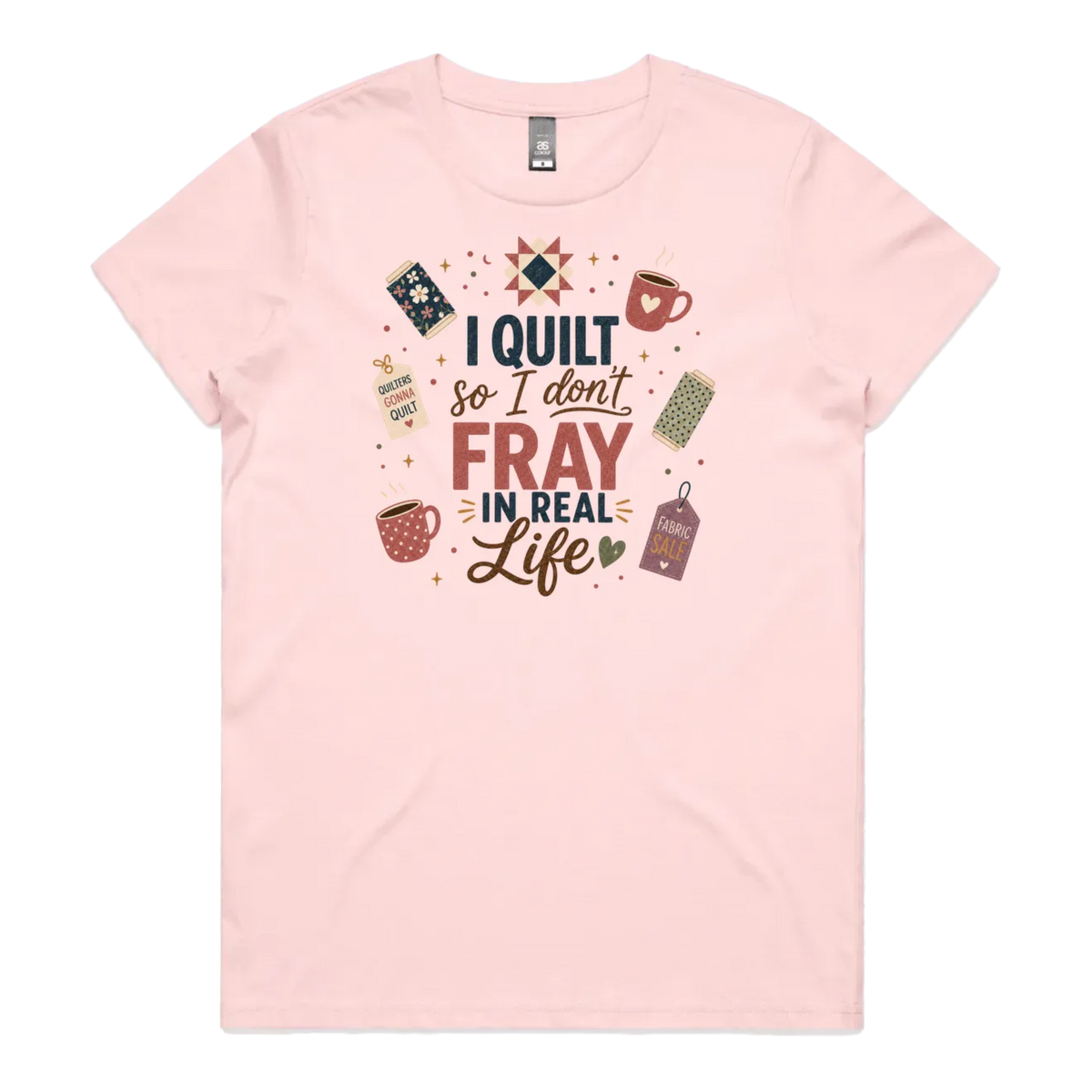 A pale pink short-sleeve crewneck t-shirt featuring a colorful graphic print on the front with quilting-themed illustrations and the text 'I Quilt So I Don't Fray in Real Life'.