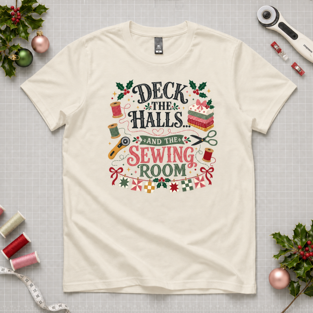 An off-white short-sleeve crewneck t-shirt featuring a colorful graphic print on the front with sewing-themed illustrations and the text 'Deck the Halls and the Sewing Room'. The shirt lays flat on a grey cutting mat with quilting tools and holly in the corners.