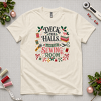 An off-white short-sleeve crewneck t-shirt featuring a colorful graphic print on the front with sewing-themed illustrations and the text 'Deck the Halls and the Sewing Room'. The shirt lays flat on a grey cutting mat with quilting tools and holly in the corners.