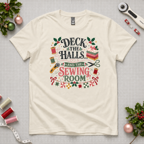 An off-white short-sleeve crewneck t-shirt featuring a colorful graphic print on the front with sewing-themed illustrations and the text 'Deck the Halls and the Sewing Room'. The shirt lays flat on a grey cutting mat with quilting tools and holly in the corners.