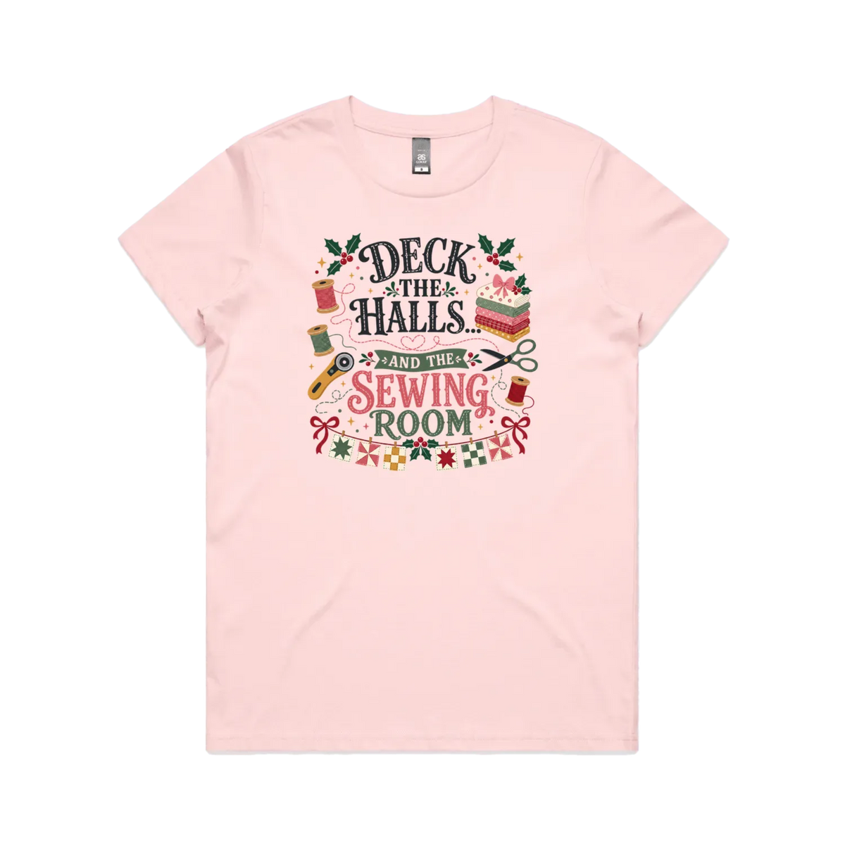 A pale pink short-sleeve crewneck t-shirt featuring a colorful graphic print on the front with sewing-themed illustrations and the text 'Deck the Halls and the Sewing Room'.