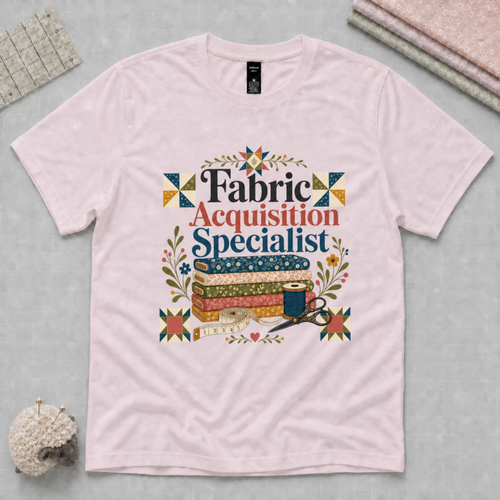 A pale mauve graphic t-shirt with the words 'Fabric Acquisition Specialist' above a stack of fabric bolts, measuring tape, thread spool and scissors.