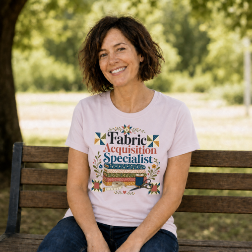 A woman with short dark hair sitting on a park bench wearing a pale mauve graphic t-shirt with the words 'Fabric Acquisition Specialist' above a stack of fabric bolts, measuring tape and scissors.