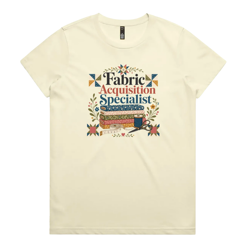 A pale butter yellow crewneck short-sleeve t-shirt with the words 'Fabric Acquisition Specialist' above a stack of fabric bolts, a measuring tape, thread spool and scissors.