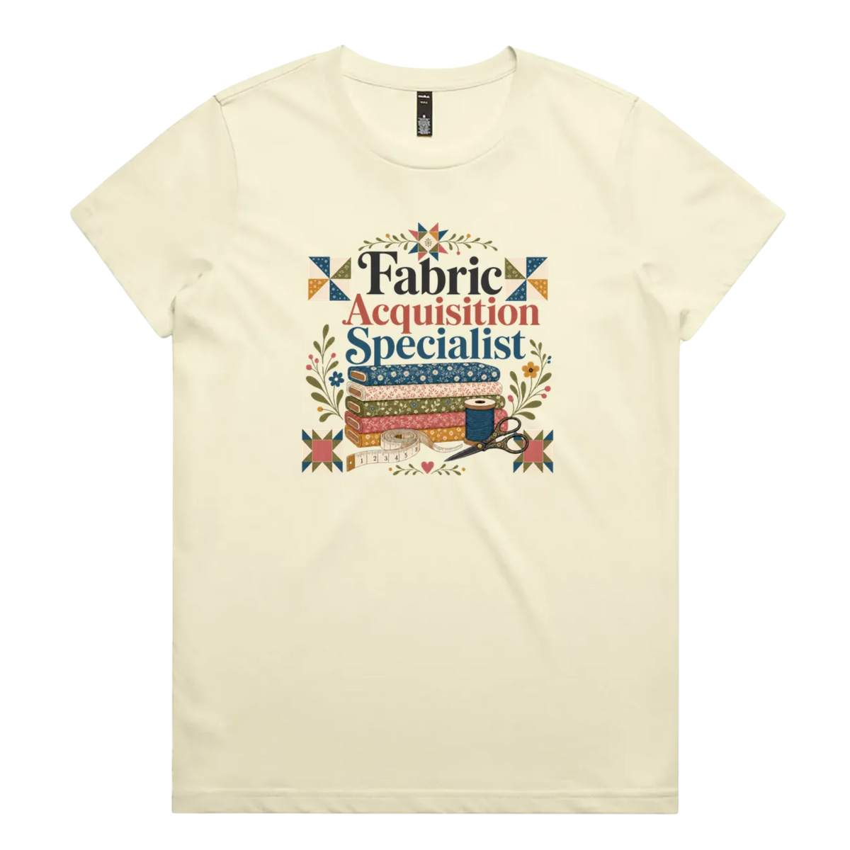 A pale butter yellow crewneck short-sleeve t-shirt with the words 'Fabric Acquisition Specialist' above a stack of fabric bolts, a measuring tape, thread spool and scissors.