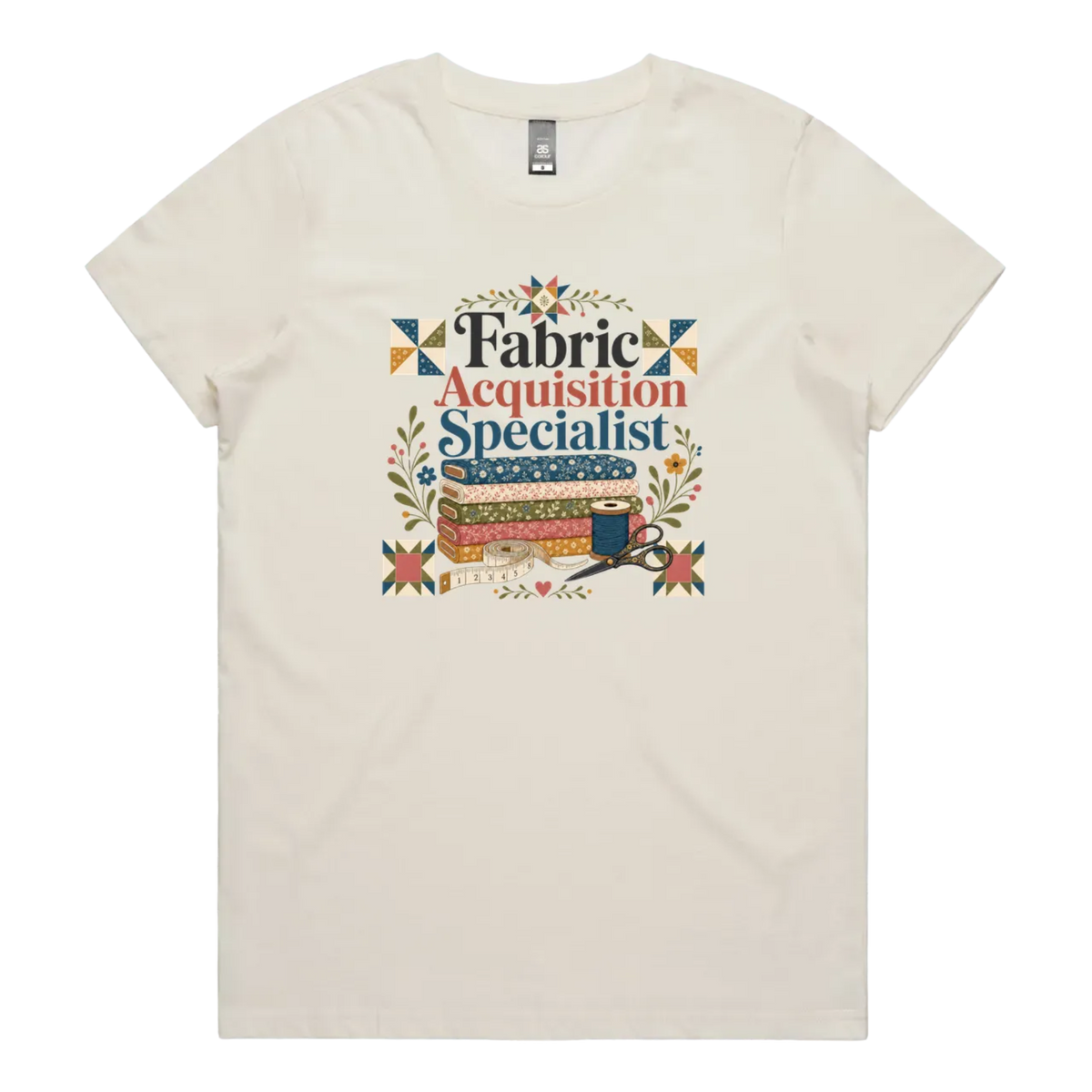 A pale cream crewneck short-sleeve t-shirt with the words 'Fabric Acquisition Specialist' above a stack of fabric bolts, a measuring tape, thread spool and scissors.