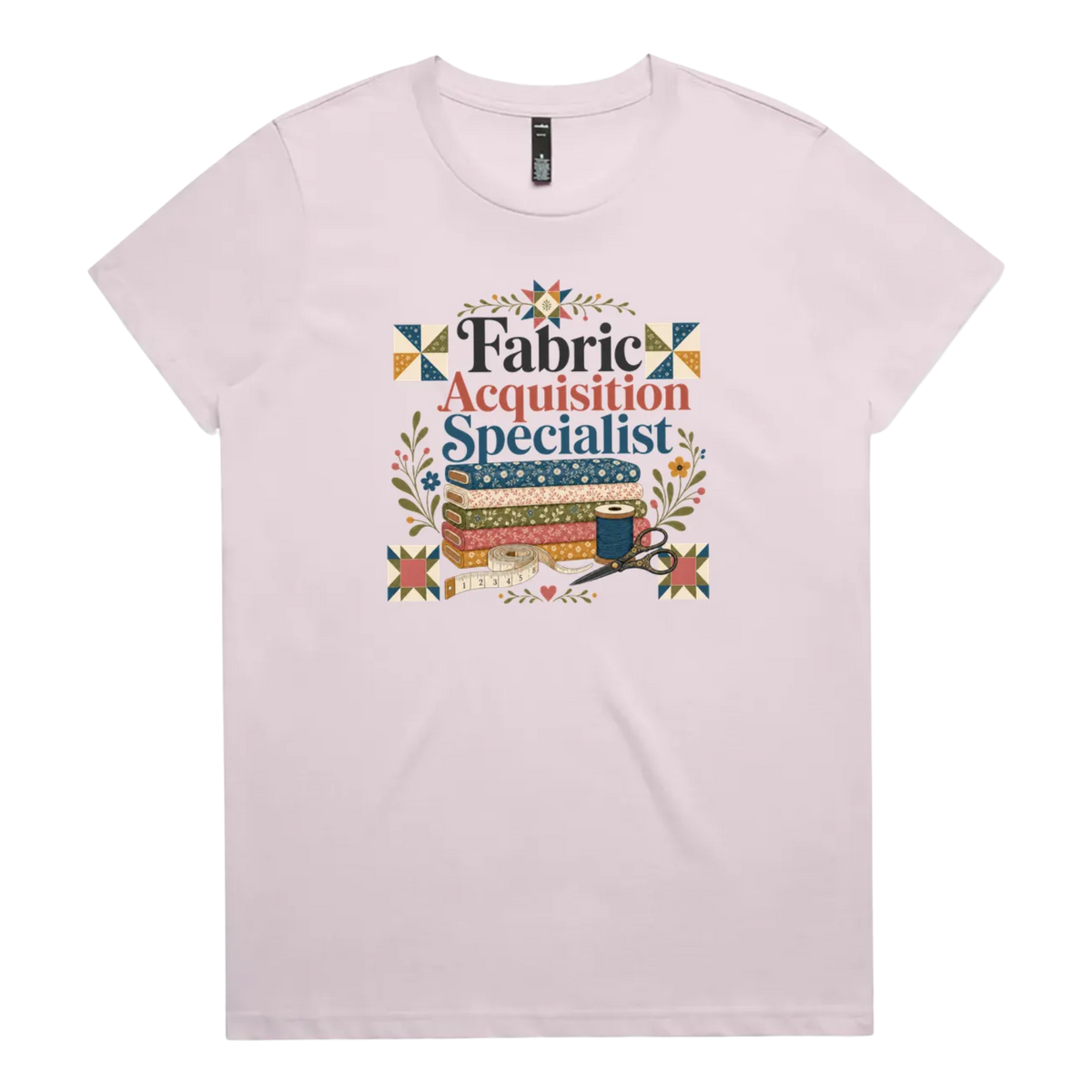 A pale mauve crewneck short-sleeve t-shirt with the words 'Fabric Acquisition Specialist' above a stack of fabric bolts, a measuring tape, thread spool and scissors.