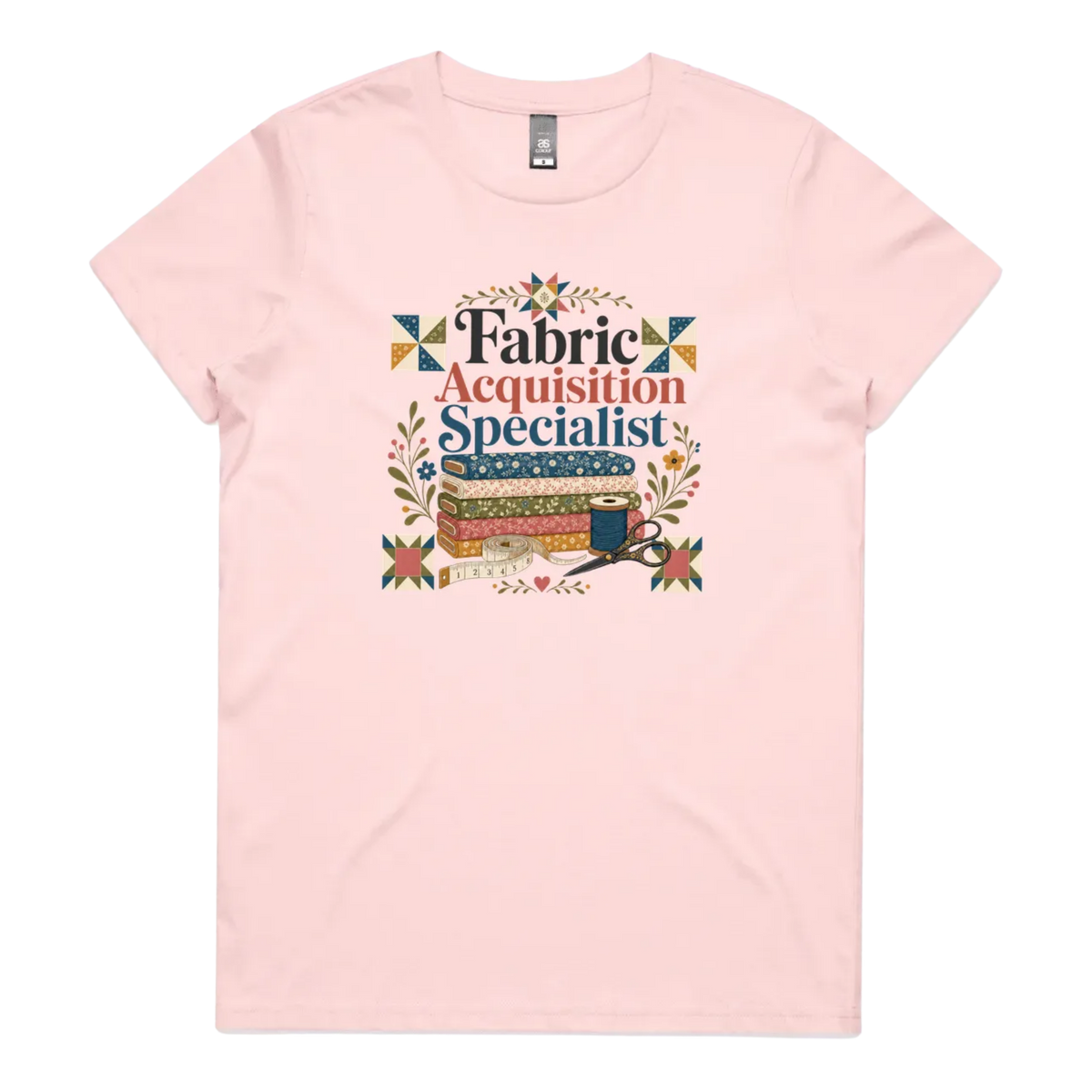 A pale pink crewneck short-sleeve t-shirt with the words 'Fabric Acquisition Specialist' above a stack of fabric bolts, a measuring tape, thread spool and scissors.