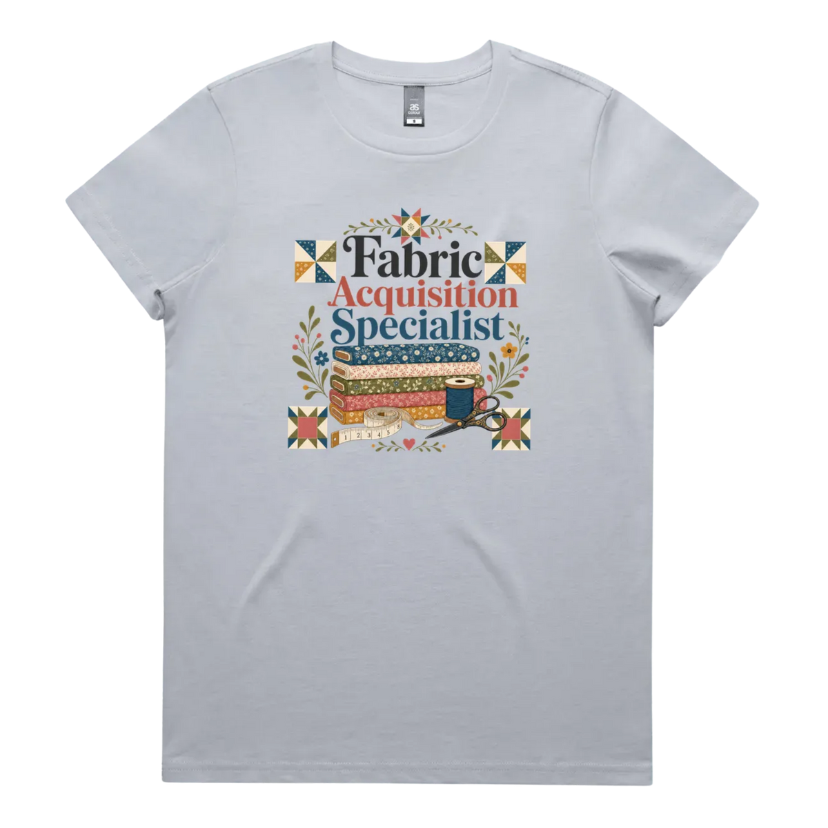 A pale powder blue crewneck short-sleeve t-shirt with the words 'Fabric Acquisition Specialist' above a stack of fabric bolts, a measuring tape, thread spool and scissors.