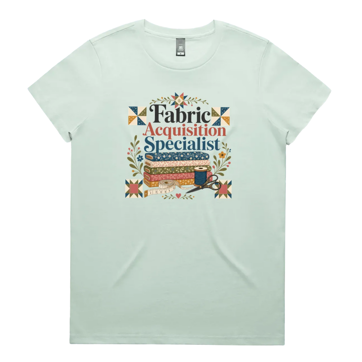 A pale seafoam green crewneck short-sleeve t-shirt with the words 'Fabric Acquisition Specialist' above a stack of fabric bolts, a measuring tape, thread spool and scissors.