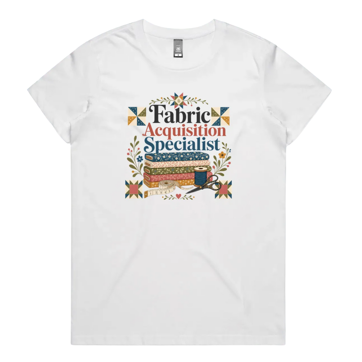 A white crewneck short-sleeve t-shirt with the words 'Fabric Acquisition Specialist' above a stack of fabric bolts, a measuring tape, thread spool and scissors.