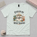 A light mint green short-sleeve t-shirt with a graphic print that reads 'Fueled by Coffee & Quilt Blocks' in black text, surrounding a vintage-style illustration of a coffee mug, thread spools, and fabric squares. The shirt is displayed flat on a light grey cutting mat with sewing props.