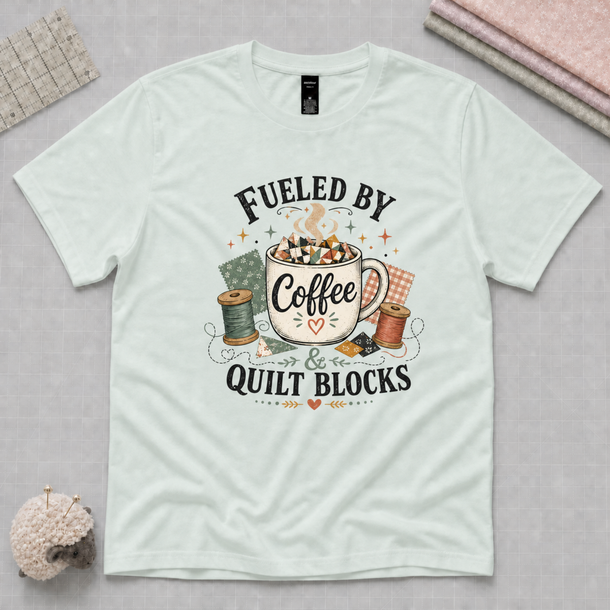 A light mint green short-sleeve t-shirt with a graphic print that reads 'Fueled by Coffee & Quilt Blocks' in black text, surrounding a vintage-style illustration of a coffee mug, thread spools, and fabric squares. The shirt is displayed flat on a light grey cutting mat with sewing props.
