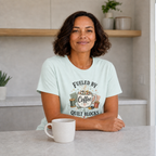 A woman with short dark curly hair sits at a kitchen bench wearing a light mint green graphic t-shirt that reads 'Fueled by Coffee & Quilt Blocks'. A white ceramic mug sits in front of her, with a modern kitchen background featuring light gray walls and wooden shelves.