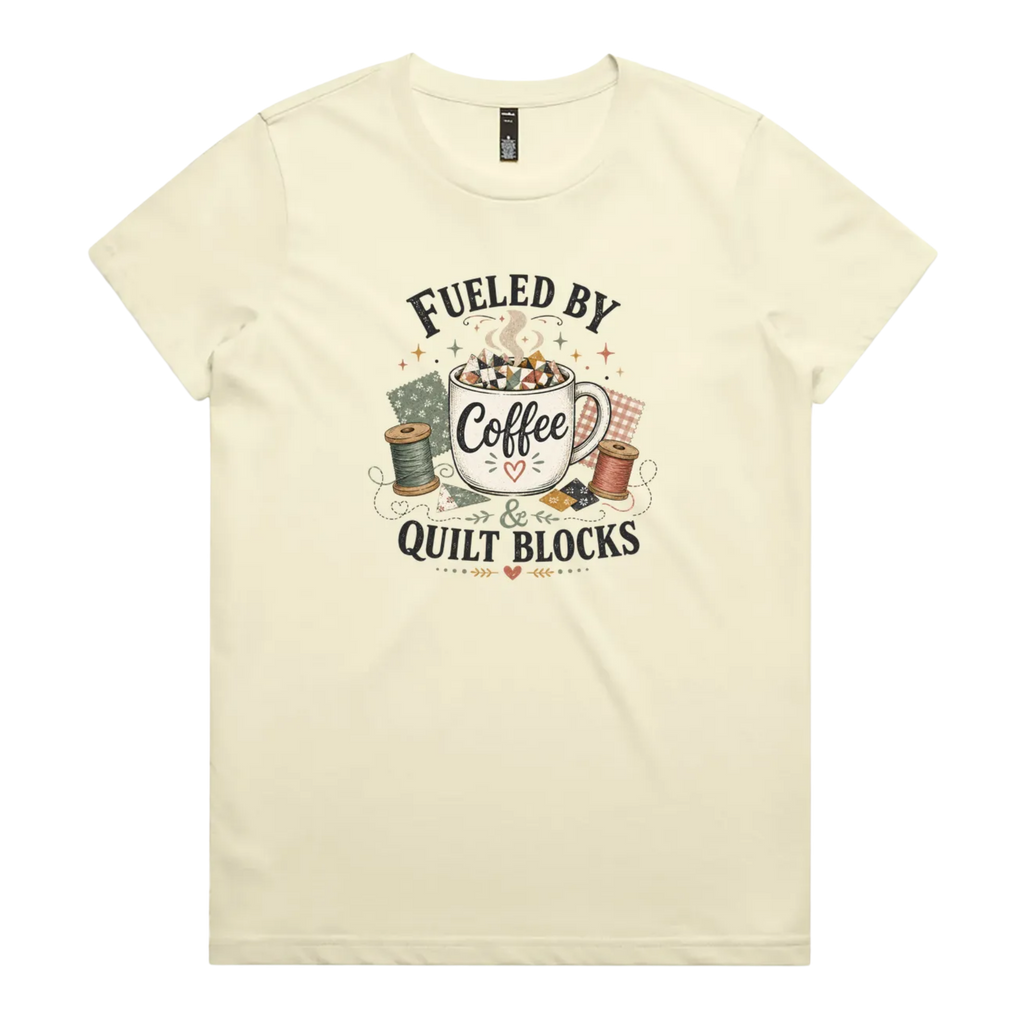 A pale butter yellow short-sleeve t-shirt with a vintage-style graphic on the front that reads 'Fueled by Coffee & Quilt Blocks' around an illustration of a coffee mug, quilting thread spools, and fabric.