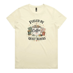 A pale butter yellow short-sleeve t-shirt with a vintage-style graphic on the front that reads 'Fueled by Coffee & Quilt Blocks' around an illustration of a coffee mug, quilting thread spools, and fabric.