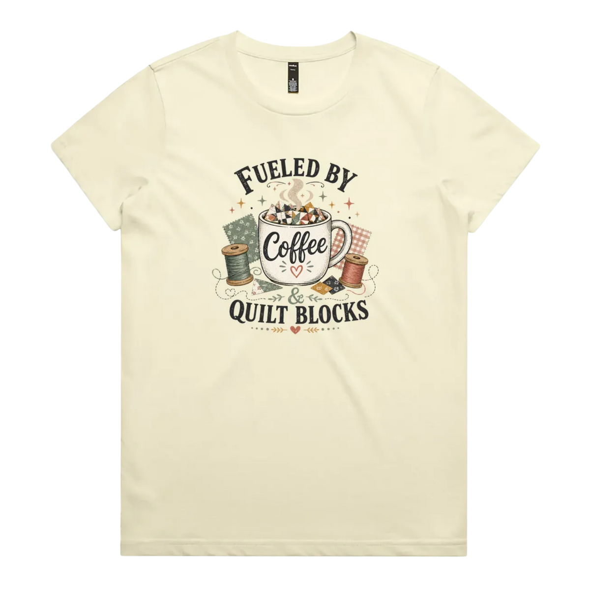 A pale butter yellow short-sleeve t-shirt with a vintage-style graphic on the front that reads 'Fueled by Coffee & Quilt Blocks' around an illustration of a coffee mug, quilting thread spools, and fabric.