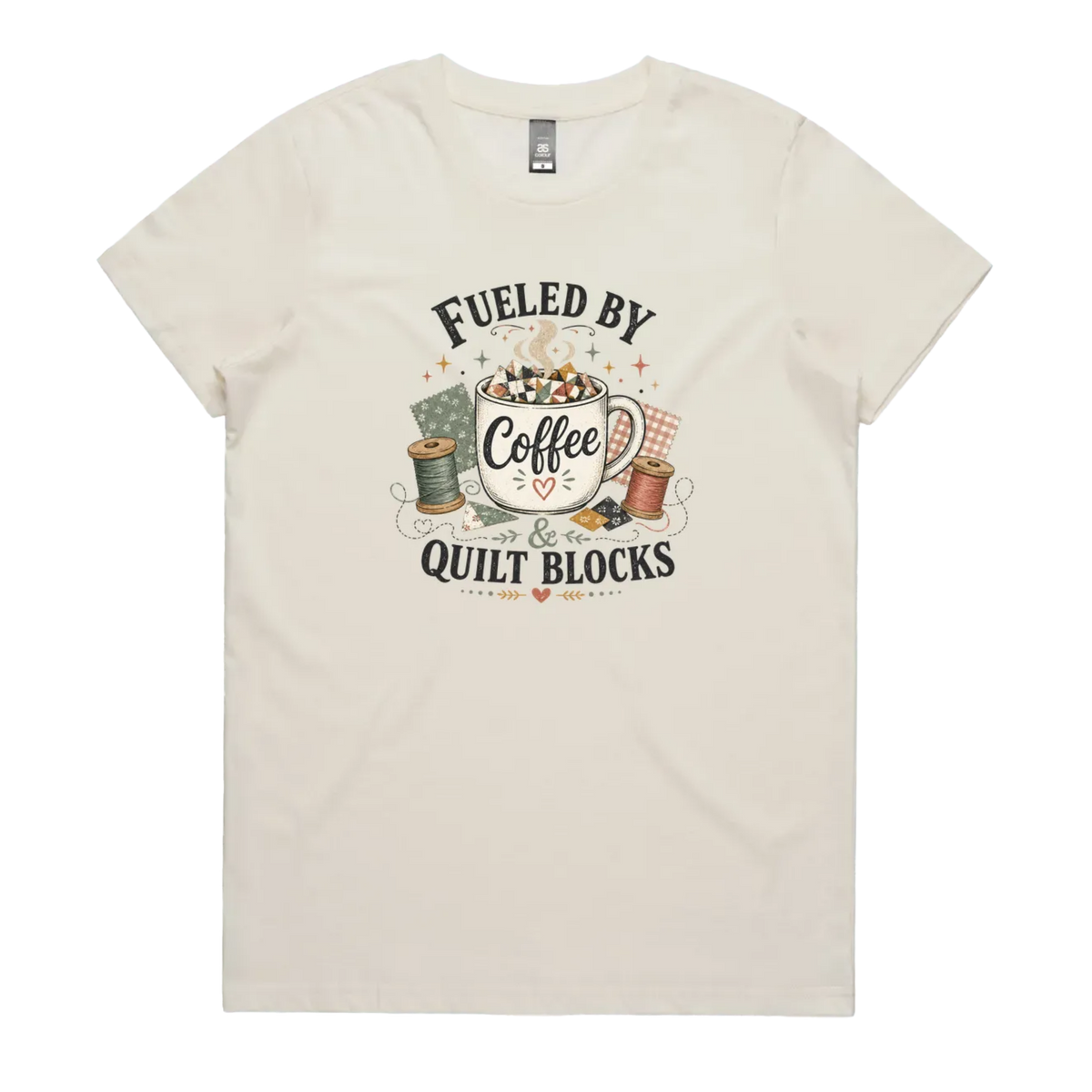 A pale cream short-sleeve t-shirt with a vintage-style graphic on the front that reads 'Fueled by Coffee & Quilt Blocks' around an illustration of a coffee mug, quilting thread spools, and fabric.