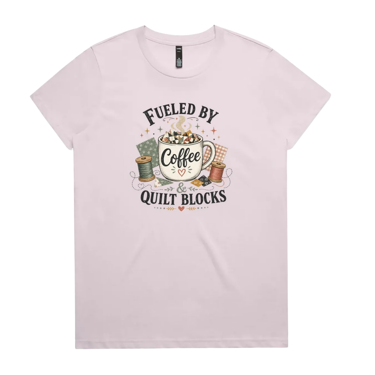 A pale mauve short-sleeve t-shirt with a vintage-style graphic on the front that reads 'Fueled by Coffee & Quilt Blocks' around an illustration of a coffee mug, quilting thread spools, and fabric.