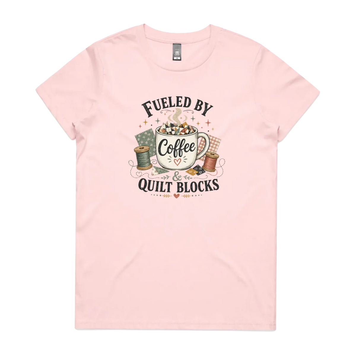 A pale pink short-sleeve t-shirt with a vintage-style graphic on the front that reads 'Fueled by Coffee & Quilt Blocks' around an illustration of a coffee mug, quilting thread spools, and fabric.