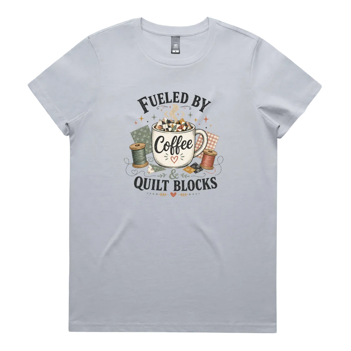 A pale powder blue short-sleeve t-shirt with a vintage-style graphic on the front that reads 'Fueled by Coffee & Quilt Blocks' around an illustration of a coffee mug, quilting thread spools, and fabric.