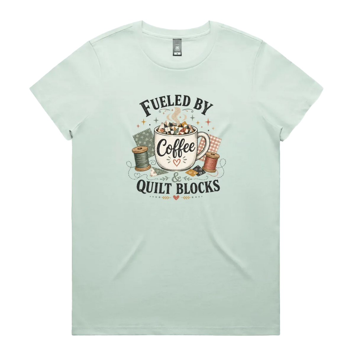A light mint green short-sleeve t-shirt with a vintage-style graphic on the front that reads 'Fueled by Coffee & Quilt Blocks' around an illustration of a coffee mug, quilting thread spools, and fabric.