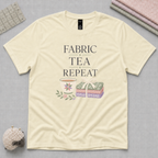 A pale butter yellow short-sleeve t-shirt with a graphic print that reads 'FABRIC, TEA, REPEAT' above an illustration of a teacup and a stack of fabric tied with a ribbon. The shirt is laid flat on a gray cutting mat with folded fabrics, quilting ruler and sheep-shaped pincushion partially visible in the corners.