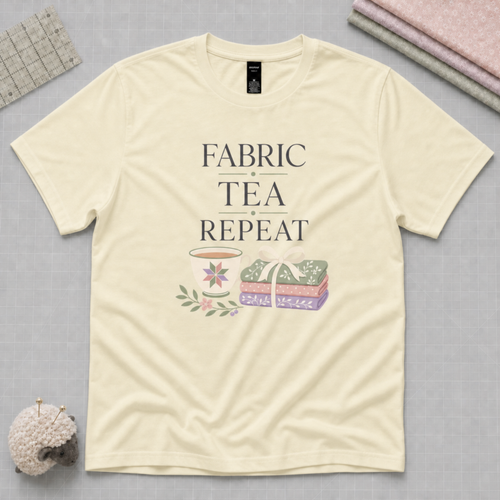A pale butter yellow short-sleeve t-shirt with a graphic print that reads 'FABRIC, TEA, REPEAT' above an illustration of a teacup and a stack of fabric tied with a ribbon. The shirt is laid flat on a gray cutting mat with folded fabrics, quilting ruler and sheep-shaped pincushion partially visible in the corners.