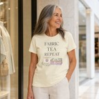 A woman with long grey hair stands in front of a shop window wearing a pale butter yellow short-sleeve t-shirt with the words "Fabric, Tea, Repeat" above an illustration of a tea cup and bundle of fabric.
