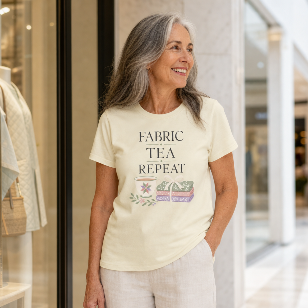 A woman with long grey hair stands in front of a shop window wearing a pale butter yellow short-sleeve t-shirt with the words "Fabric, Tea, Repeat" above an illustration of a tea cup and bundle of fabric.