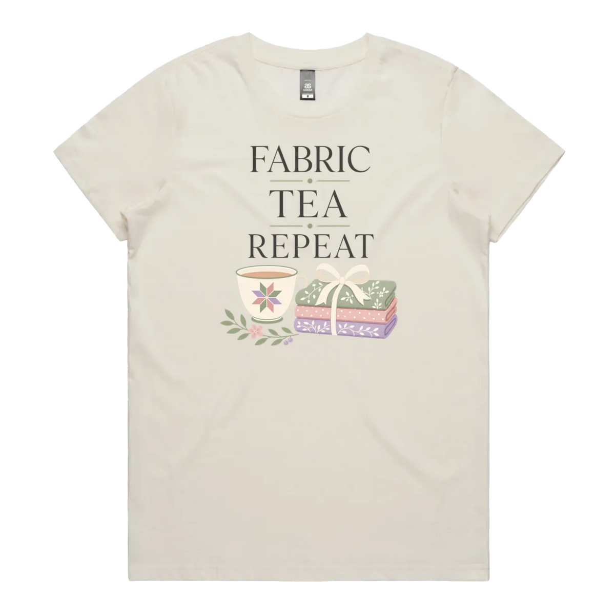 An off-white short-sleeve crewneck t-shirt featuring a graphic print that reads 'FABRIC, TEA, REPEAT' above an illustration of a teacup, floral details, and patterned fabric stack.