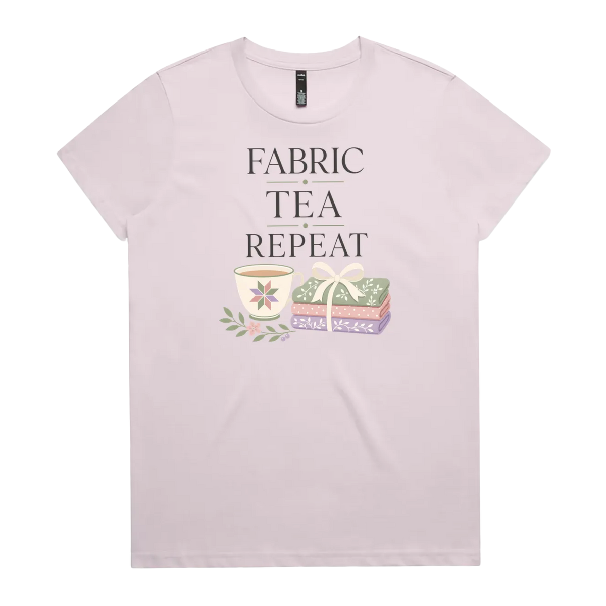 A pale mauve short-sleeve crewneck t-shirt featuring a graphic print that reads 'FABRIC, TEA, REPEAT' above an illustration of a teacup, floral details, and patterned fabric stack.