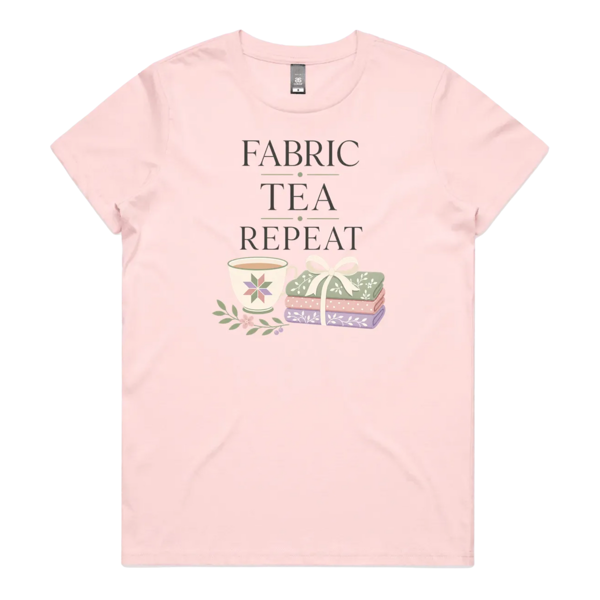 A pale pink short-sleeve crewneck t-shirt featuring a graphic print that reads 'FABRIC, TEA, REPEAT' above an illustration of a teacup, floral details, and patterned fabric stack.