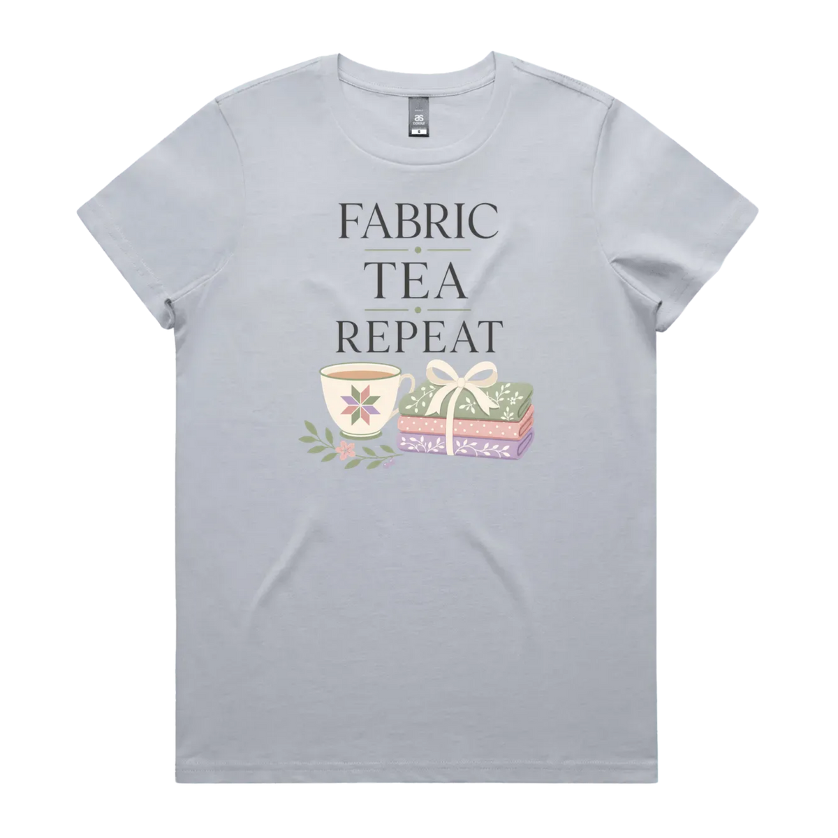 A powder blue short-sleeve crewneck t-shirt featuring a graphic print that reads 'FABRIC, TEA, REPEAT' above an illustration of a teacup, floral details, and patterned fabric stack.