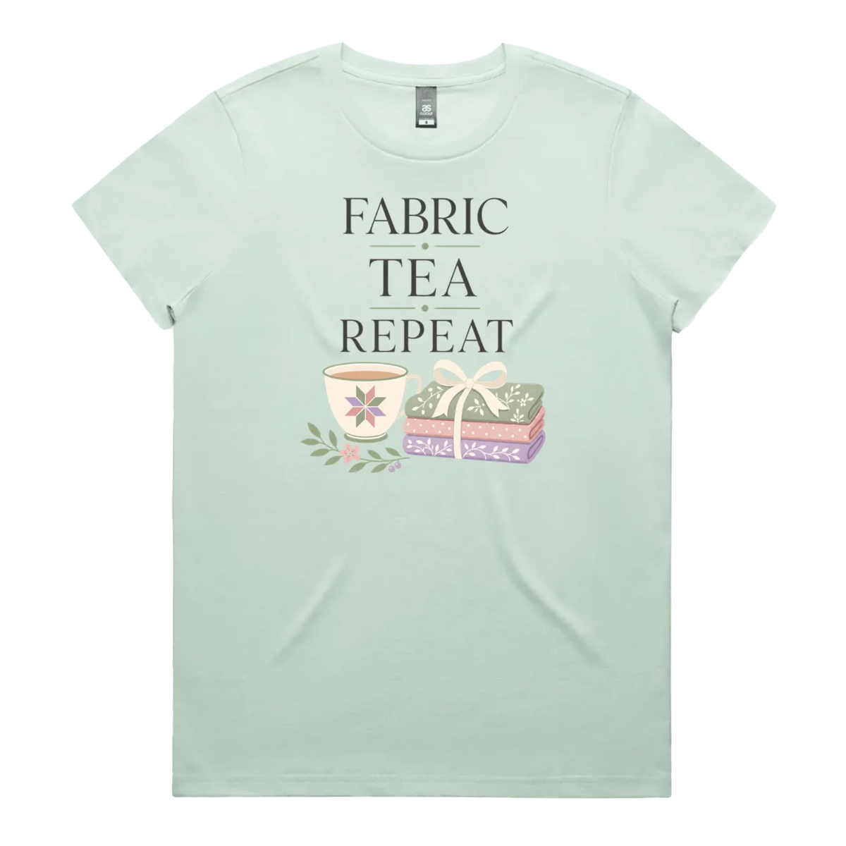 A pale mint green short-sleeve crewneck t-shirt featuring a graphic print that reads 'FABRIC, TEA, REPEAT' above an illustration of a teacup, floral details, and patterned fabric stack.