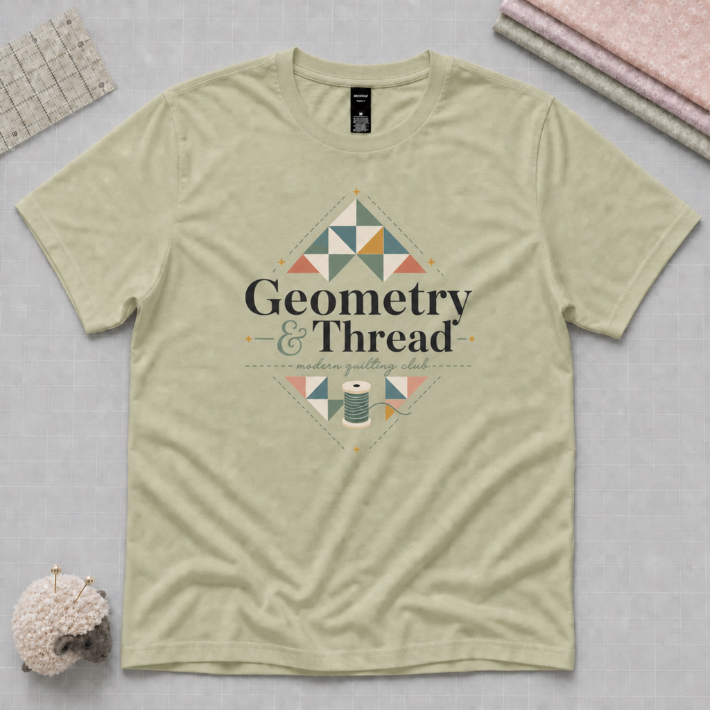 A pistachio green short-sleeve t-shirt laying flat on a pale grey cutting mat withe quilting tools in the corners. The t-shirt features a quilt-themed graphic with the words 'Geometry & Thread' surrounded by patchwork triangles.
