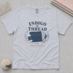 A powder blue crewneck t-shirt  featuring a navy blue and blue-gray graphic print that reads 'INDIGO & THREAD', surrounded by sashiko-themed illustrations. The shirt lays flat on a pale grey cutting mat with sewing props in the corners.