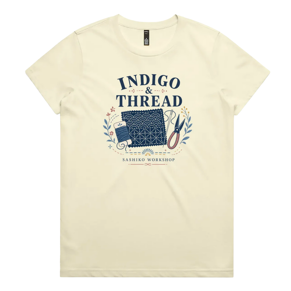 A pale butter yellow short-sleeve crewneck t-shirt featuring a navy blue and multicolor graphic on the front that reads 'INDIGO & THREAD' with a spool of thread, sashiko fabric swatch, scissors, and 'SASHIKO WORKSHOP' printed below.