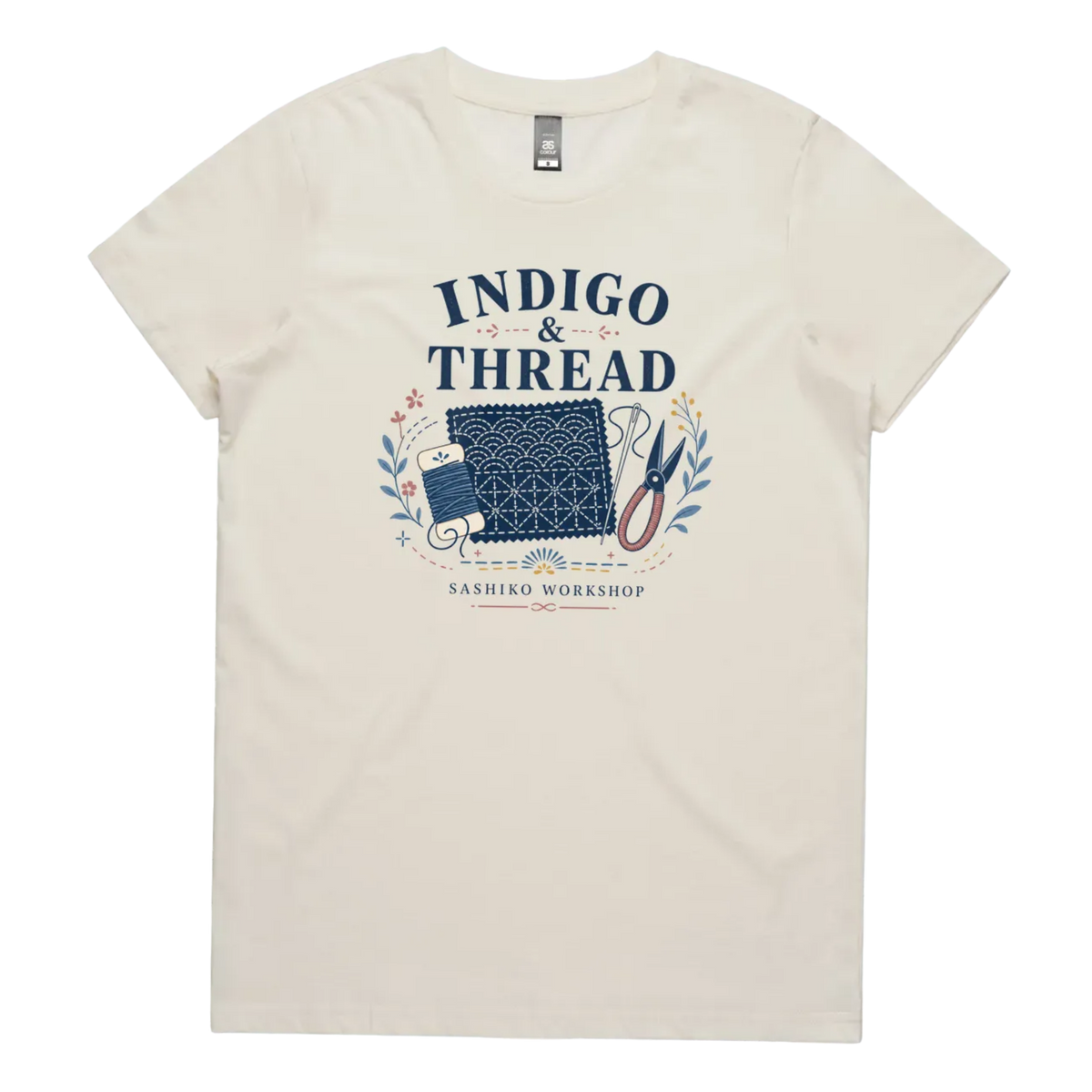 A pale cream short-sleeve crewneck t-shirt featuring a navy blue and multicolor graphic on the front that reads 'INDIGO & THREAD' with a spool of thread, sashiko fabric swatch, scissors, and 'SASHIKO WORKSHOP' printed below.
