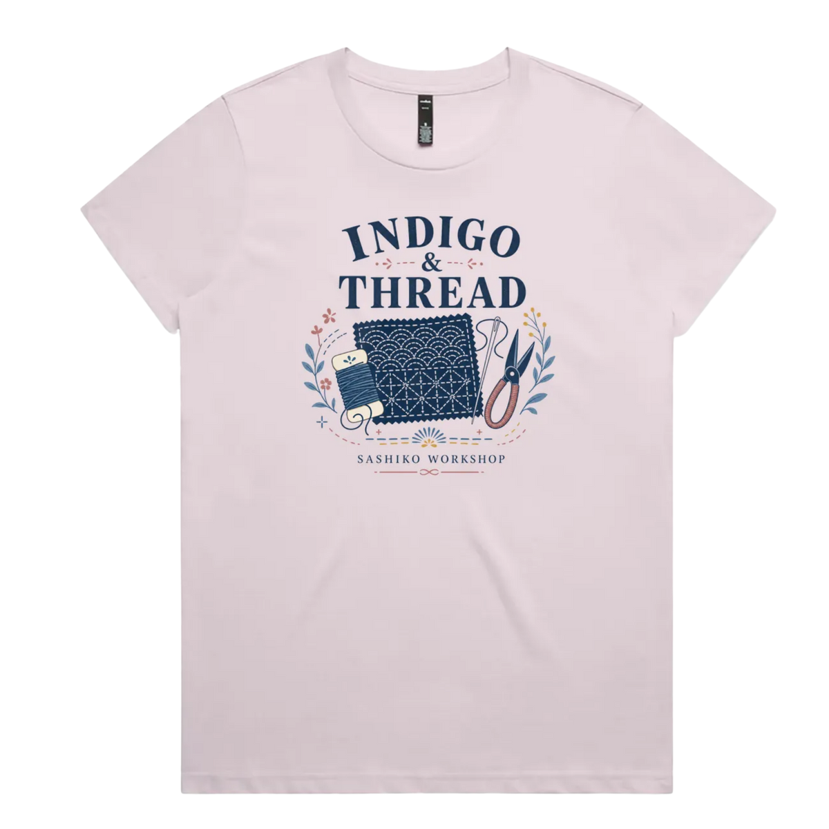 A pale mauve short-sleeve crewneck t-shirt featuring a navy blue and multicolor graphic on the front that reads 'INDIGO & THREAD' with a spool of thread, sashiko fabric swatch, scissors, and 'SASHIKO WORKSHOP' printed below.