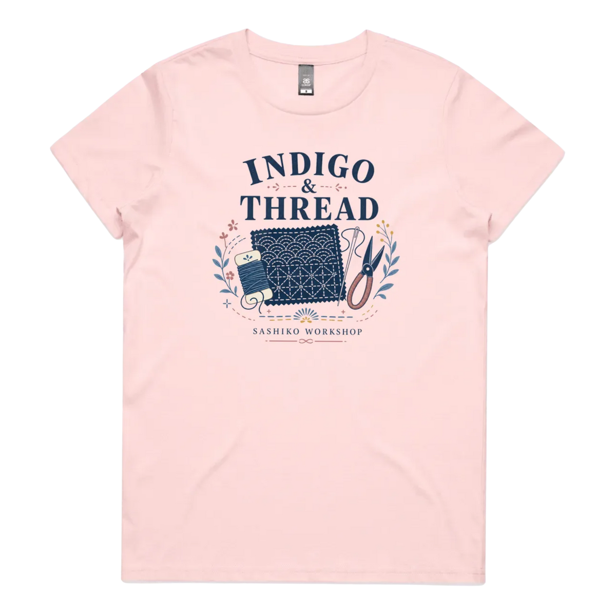 A pale pink short-sleeve crewneck t-shirt featuring a navy blue and multicolor graphic on the front that reads 'INDIGO & THREAD' with a spool of thread, sashiko fabric swatch, scissors, and 'SASHIKO WORKSHOP' printed below.