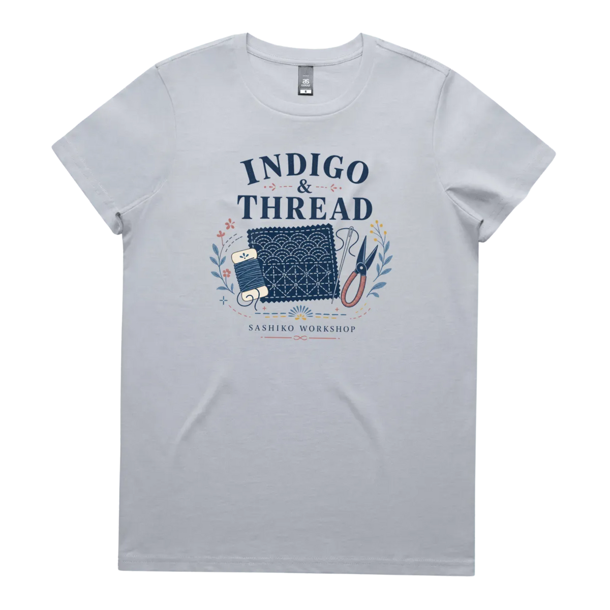 A pale powder blue short-sleeve crewneck t-shirt featuring a navy blue and multicolor graphic on the front that reads 'INDIGO & THREAD' with a spool of thread, sashiko fabric swatch, scissors, and 'SASHIKO WORKSHOP' printed below.