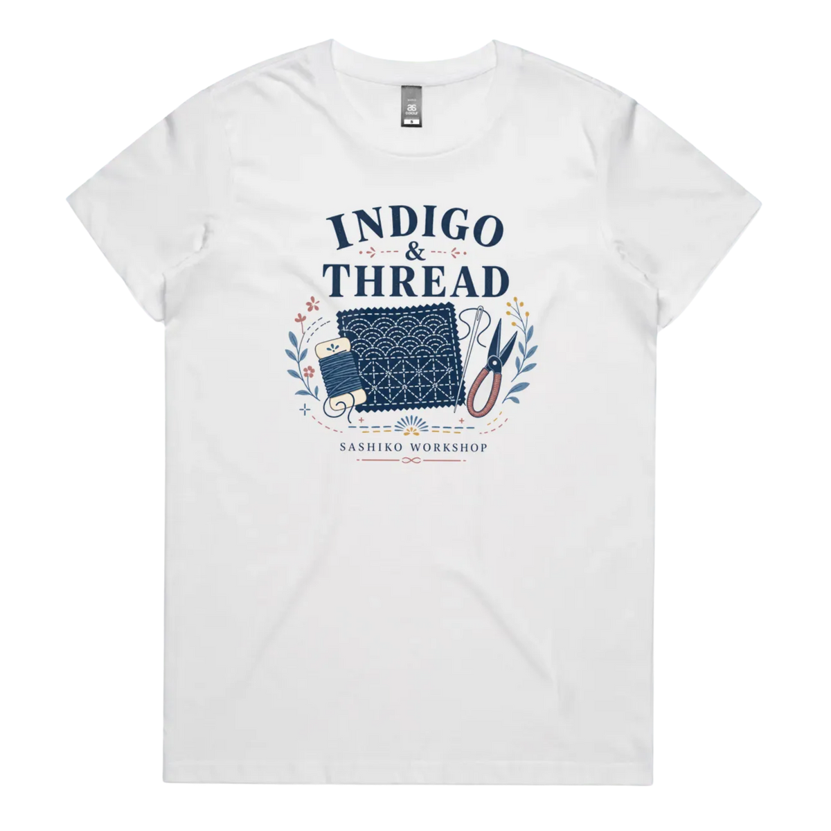 A white short-sleeve crewneck t-shirt featuring a navy blue and multicolor graphic on the front that reads 'INDIGO & THREAD' with a spool of thread, sashiko fabric swatch, scissors, and 'SASHIKO WORKSHOP' printed below.