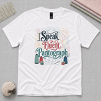 A white short-sleeve graphic T-shirt laid flat on a gray surface, featuring the phrase 'I Speak Fluent Pantograph' in decorative multicolor lettering with sewing-themed illustrations. Styled flat-lay props include a quilting ruler, folded fabric stack, and a small pincushion around the shirt.