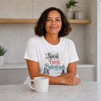 A woman with short dark curly hair sits at a light gray table in a bright kitchen, wearing a white graphic T-shirt that reads 'I Speak Fluent Pantograph' with decorative lettering and illustrations. A white mug sits in front of her, and shelves with plants are blurred in the background.