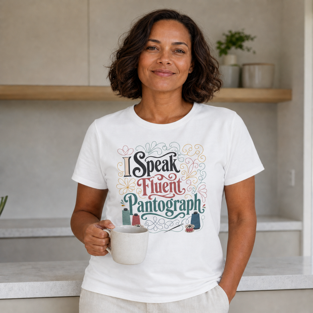 A woman with short dark hair stands in a kitchen setting, wearing a white graphic T-shirt that reads 'I Speak Fluent Pantograph' in decorative multicolor lettering, and holding a white mug.