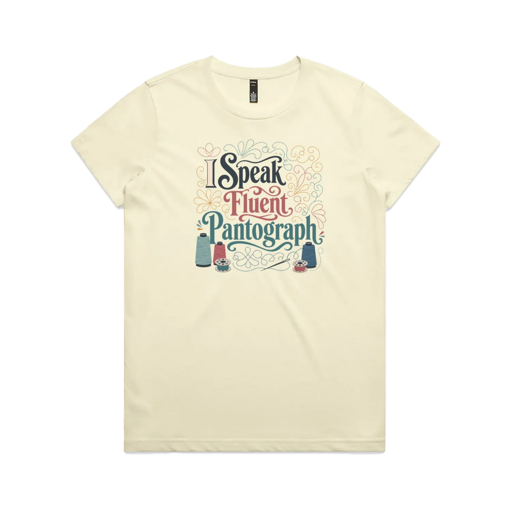 A pale butter yellow graphic short-sleeve t-shirt with the words 'I Speak Fluent Pantrograph' surrounded by thread spools, bobbins, a needle and swirling stitche lines in multi colours.