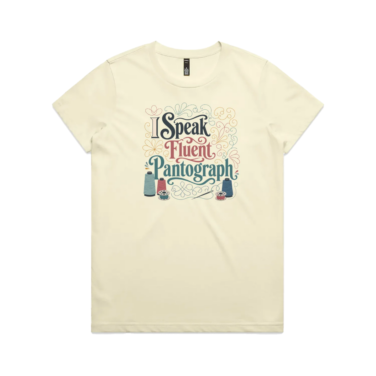 A pale butter yellow graphic short-sleeve t-shirt with the words 'I Speak Fluent Pantrograph' surrounded by thread spools, bobbins, a needle and swirling stitche lines in multi colours.