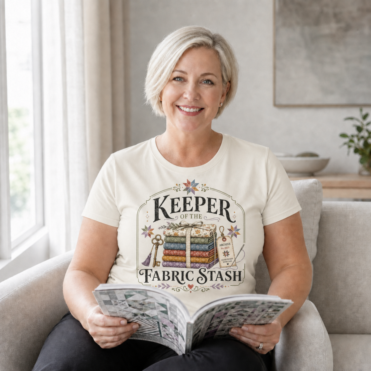 A woman with short blond hair sitting on a beige lounge holding an open book. She is wearing a cream coloured graphic t-shirt with the words 'Keeper of the Fabric Stash' with a stack of folded fabric, a key, needle, archive tag and small stars.